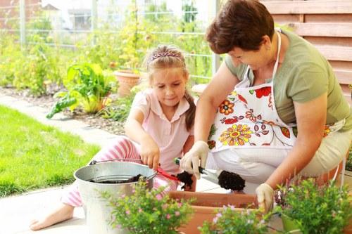The image depicts a young girl and an adult woman working together in an outdoor garden space, likely during daytime with natural light. They are seated on a paved patio area adjacent to a lush, well-maintained lawn. The girl, with light brown hair tied back, is wearing a light pink shirt and pink and white striped pants, and she is actively handling a small gardening trowel. The woman, wearing a light green top and a white apron with a floral pattern, is assisting her with gardening tasks. In front of them are several vibrant green plants with small purple flowers placed in terracotta pots, along with garden tools and a metal bucket. The background features a wooden garden fence, a variety of green shrubs, and some trees, suggesting a private backyard or front garden area in Muswell Hill. The scene captures a moment of outdoor gardening activity, emphasizing garden cultivation, planting, and family engagement in maintenance, aligning with professional gardening services provided by Landscapers Muswell Hill. The weather appears clear, contributing to the bright and lively atmosphere of the garden environment.