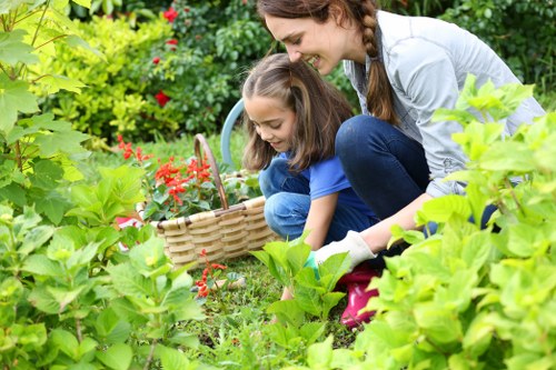 A woman and a young girl are gardening together in a lush, well-maintained outdoor space, surrounded by various green shrubs and plants. The girl is holding a woven basket and appears to be picking flowers or berries, while the woman is kneeling beside her, smiling and assisting. In the background, there are flowering plants with red blooms, and the garden features a mix of dense foliage, ground cover, and possibly a small paved area. The scene suggests a pleasant, sunny day with natural lighting highlighting the vibrant greens and colorful flowers typical of a cared-for lawn and garden environment in Muswell Hill. This outdoor space showcases an inviting and organized garden layout, with an emphasis on natural textures and seasonal plants, aligning with a professional landscaping and gardening service approach, as provided by Landscapers Muswell Hill.