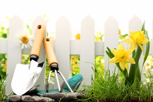 The image depicts a section of a well-maintained garden with a white picket fence in the background, typical of outdoor spaces in Muswell Hill. In the foreground, there is a patch of lush, green grass with a dense, healthy appearance, bordered by a flower bed containing vibrant yellow daffodils and some white daisies. The flower bed is positioned adjacent to paved paving stones that provide a clean, organized border between the lawn and planting area. Among the flowers, garden hand tools such as a small trowel, a hand fork, and pruning shears are neatly arranged, suggesting recent gardening activity. Bright sunlight illuminates the space, creating natural shadows and highlighting the fresh growth and vivid colours of the flowers. The overall scene reflects a tidy, inviting outdoor area suitable for ongoing gardening and landscaping work, typical of residential gardens in the Muswell Hill area, which fits within the scope of services offered by Landscapers Muswell Hill for planting and outdoor maintenance. The environment appears to be a private garden space prepared for seasonal planting or ongoing care, emphasizing natural textures and peaceful outdoor aesthetics.