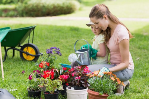 In a well-maintained garden in Muswell Hill, a young woman and a girl are engaged in gardening activities, tending to an array of potted flowering plants with vibrant red, purple, and orange blooms situated on a lush green lawn. The garden features a neatly trimmed hedge in the background, along with a paved pathway. A wheelbarrow with a green body and black handles, and yellow wheels, is positioned nearby, indicating ongoing outdoor landscaping or garden maintenance. The scene is lit by natural daylight under clear weather conditions, highlighting the natural tones of the grass and plant foliage. The garden appears to be part of a landscaped yard with a combination of grass, flower beds, and functional garden tools, contributing to a healthy outdoor environment suitable for outdoor gardening services offered by Landscapers Muswell Hill. This image exemplifies the care and attention involved in general garden upkeep, lawn care, and flower bed maintenance typical of professional gardening projects in the Muswell Hill area.