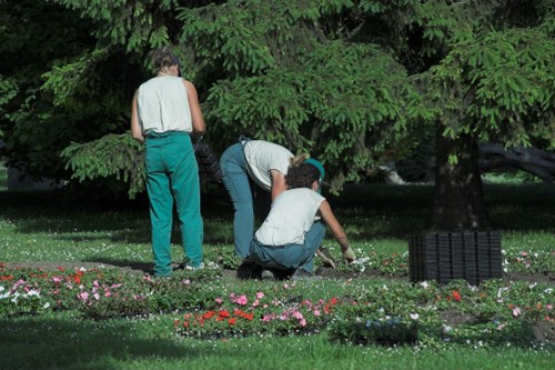 In a lush garden setting, two individuals are engaged in planting or tending to flower beds bordered with small, colourful blossoms, while a third person stands nearby under a large, leafy tree providing shade. The garden features neatly maintained grass with a mix of vibrant flower patches and dense greenery, including mature trees and hedges that form a natural boundary. The ground is a combination of lush grass, soil, and flower borders, with mature trees in the background giving a sense of enclosure. The scene captures a peaceful outdoor environment typical of suburban gardens in Muswell Hill, with natural light illuminating the space under a partly cloudy sky. The individuals are wearing casual gardening attire, indicative of outdoor maintenance work that a professional landscaping service such as Landscapers Muswell Hill might undertake to keep residential gardens inviting and well-kept, especially in suburban London areas with similar greenery.