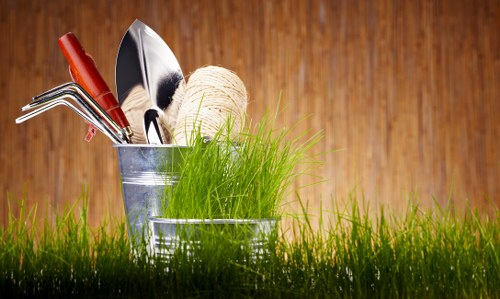 A close-up of a gardening scene featuring a small, metal container holding various gardening tools, including a trowel with a wooden handle, a pair of pruning shears, and a small rake, all arranged among vibrant green grass at the foreground. Behind the tools, there is a ball of natural twine, slightly unwound. The background consists of a warm-toned wooden surface, possibly a garden shed or fencing, providing a rustic backdrop. The image highlights the tools and materials used in garden maintenance and planting tasks, suitable for gardening and landscaping services such as those offered by Landscapers Muswell Hill in the Muswell Hill area. The composition emphasizes readiness for outdoor gardening activities, with natural lighting suggesting a clear, daylight environment, making it ideal for illustrating gardening service pages related to planting and tool use in local outdoor spaces.