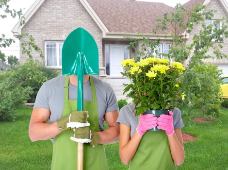 A man and a woman standing in a front garden of a residential property, both wearing green gardening aprons and pink and green gloves. The man is holding a blue shovel upright, with his face covered by a large, shiny green gardening helmet. The woman is holding a pink pot overflowing with bright yellow flowering plants, partially obscuring her face. The background features a well-maintained lawn, a paved pathway, and a brick house with white-framed windows and a gabled roof, surrounded by small trees and shrubs. The scene appears to be set on a bright, partly cloudy day in a suburban neighbourhood, highlighting outdoor gardening activities typical of local landscape improvement services in Muswell Hill.