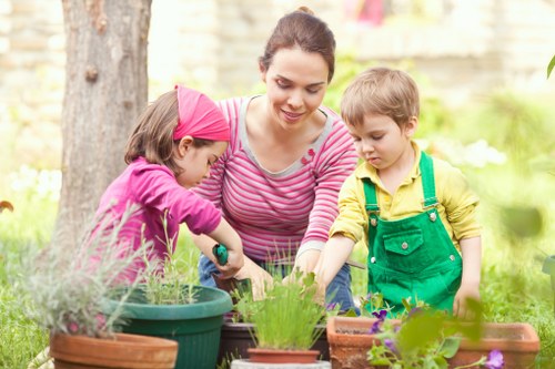 The image depicts a woman accompanied by two children engaging in outdoor gardening activities in a garden setting, with lush green grass and mature trees in the background. The woman is kneeling and smiling as she assists the children in planting or tending to potted plants and flower beds arranged on the ground. One child, a girl wearing a pink headband and jacket, is reaching into a green plant pot, while a young boy in a yellow shirt with green overalls stands nearby, observing or participating in the gardening. The garden features a mix of flower containers, some with blooming flowers and others with emerging or small plants, set on soil and paving surfaces. The environment appears bright with natural daylight, suggesting a clear day, and the overall scene conveys a peaceful, well-maintained outdoor space suitable for family gardening activities, reflecting typical gardening services offered by companies like Landscapers Muswell Hill in the local area of Muswell Hill, London.