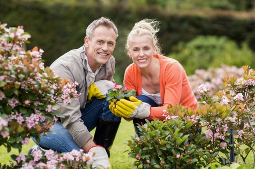 A man and woman in casual gardening attire, including yellow gardening gloves, are kneeling together in a well-maintained front garden in Muswell Hill. They are surrounded by lush, flowering shrubs with pink and white blossoms, and are smiling at the camera. The garden features a neatly trimmed hedge, blooming plants, and a vibrant green lawn, with trees and a blurred background indicating a residential outdoor space. The scene is set in natural daylight with overcast conditions, highlighting the healthy plants and the joyful outdoor activity. This outdoor environment illustrates typical gardening work performed by professionals such as those from Landscapers Muswell Hill, specializing in lawn care, hedge trimming, and garden maintenance services, creating a vibrant and inviting garden area in North London.